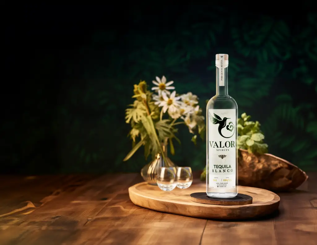 Image of Valor additive-free Blanco Tequila, stands with the hummingbird logo on a wood board, with flowers in the background